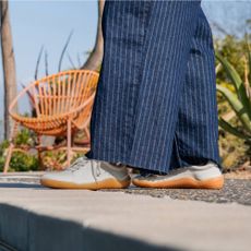 A low-down photo of someone standing on a concrete path wearing blue pinstriped trousers and white Vivobarefoot trainers with a wicker chair and green plants in the background