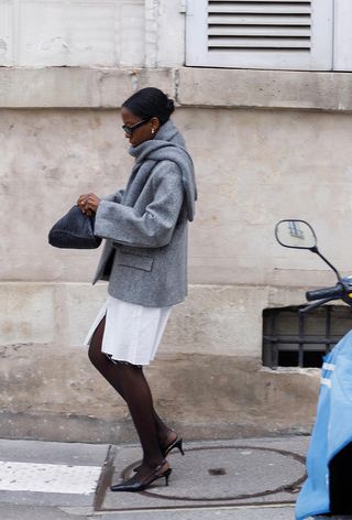 A cute work winter outfit is shown in a photo of a woman walking on the sidewalk in Paris wearing a gray cardigan over a gray wool blazer styled with a white denim skirt, black tights, black pumps, and a black clutch bag