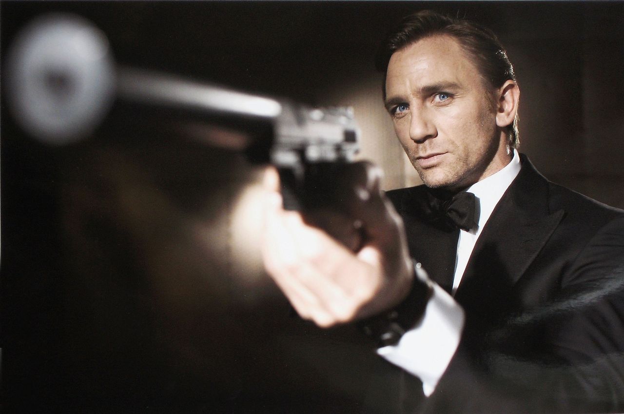 Actor Daniel Craig poses as James Bond, wearing a tuxedo, pointing a gun towards the camera