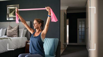 Woman sitting down on chair with resistance bands raised above her head in shoulder press as part of a chair workout