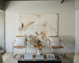 Abstract white art piece with washed out flecks of earthy colors and two wicker indoor chairs positioned in front of coffee table