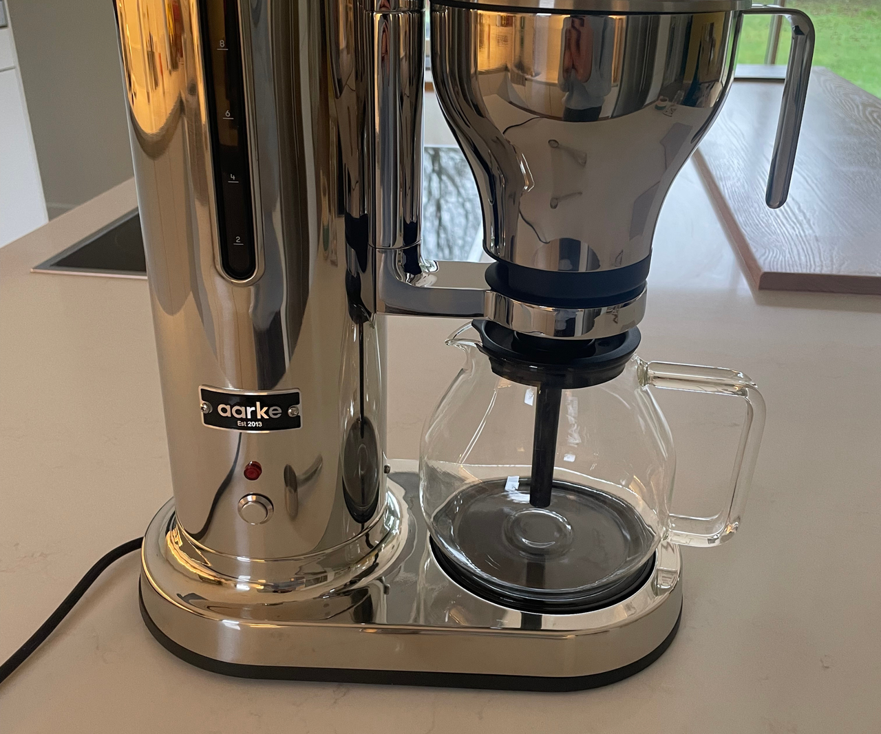 A tall stainless steel Aarke Drip Coffee Maker with a glass coffee pot on a white kitchen counter.