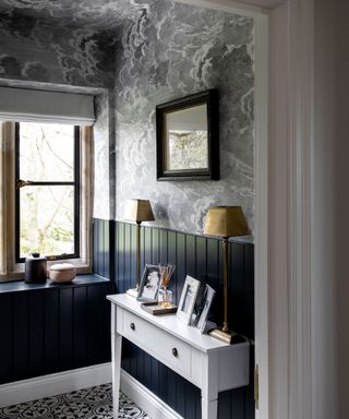 Blue and white hallway with gray cloud print wallpaper on ceiling and upper half of wall, dark blue wooden wall paneling, geometric white and blue floor tiles, white wooden console table, decorated with two matching table lamps, pictures, artwork mounted above in wooden frame, window with large shelf, decorated with ornaments