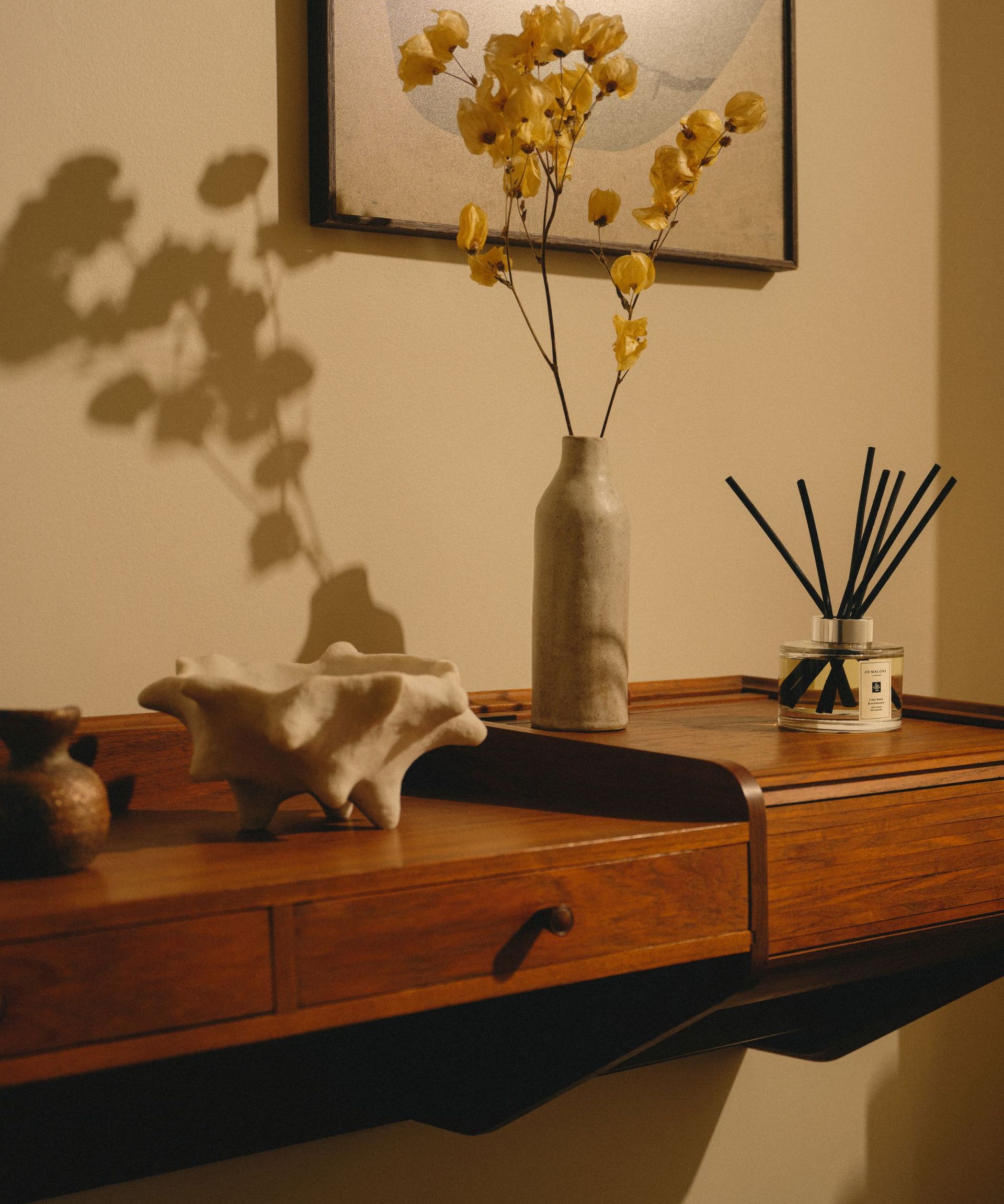 A Jo Malone reed diffuser with black reeds on a wooden countertop with a white shell and a white vase filled with yellow dried flowers. Above is a framed picture in a black frame.