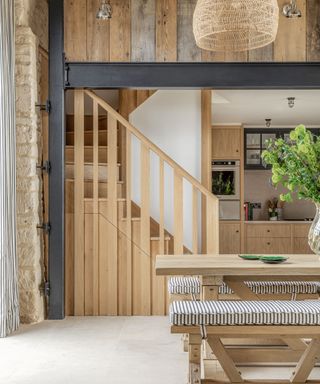 Large, farmhouse style dining room, rustic wooden beams and sleek, black metal beam, large wooden dining table with bench with seat cushion, looking onto wooden stairs and kitchen