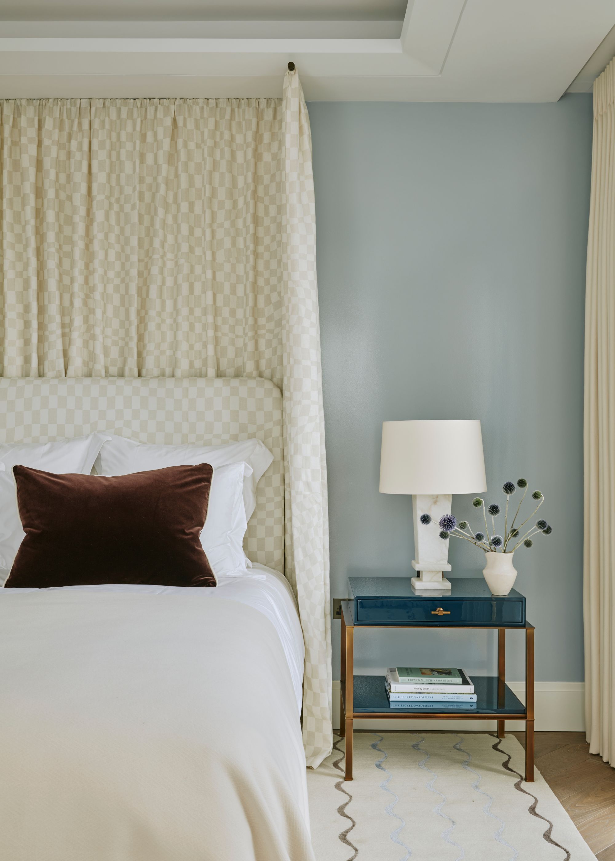 A calming bedroom with walls painted a light shade of blue, a canopy bed with a patterned neutral fabric, light neutral bedding with a chocolate brown cushion, and a blue nightstand with a white lamp.