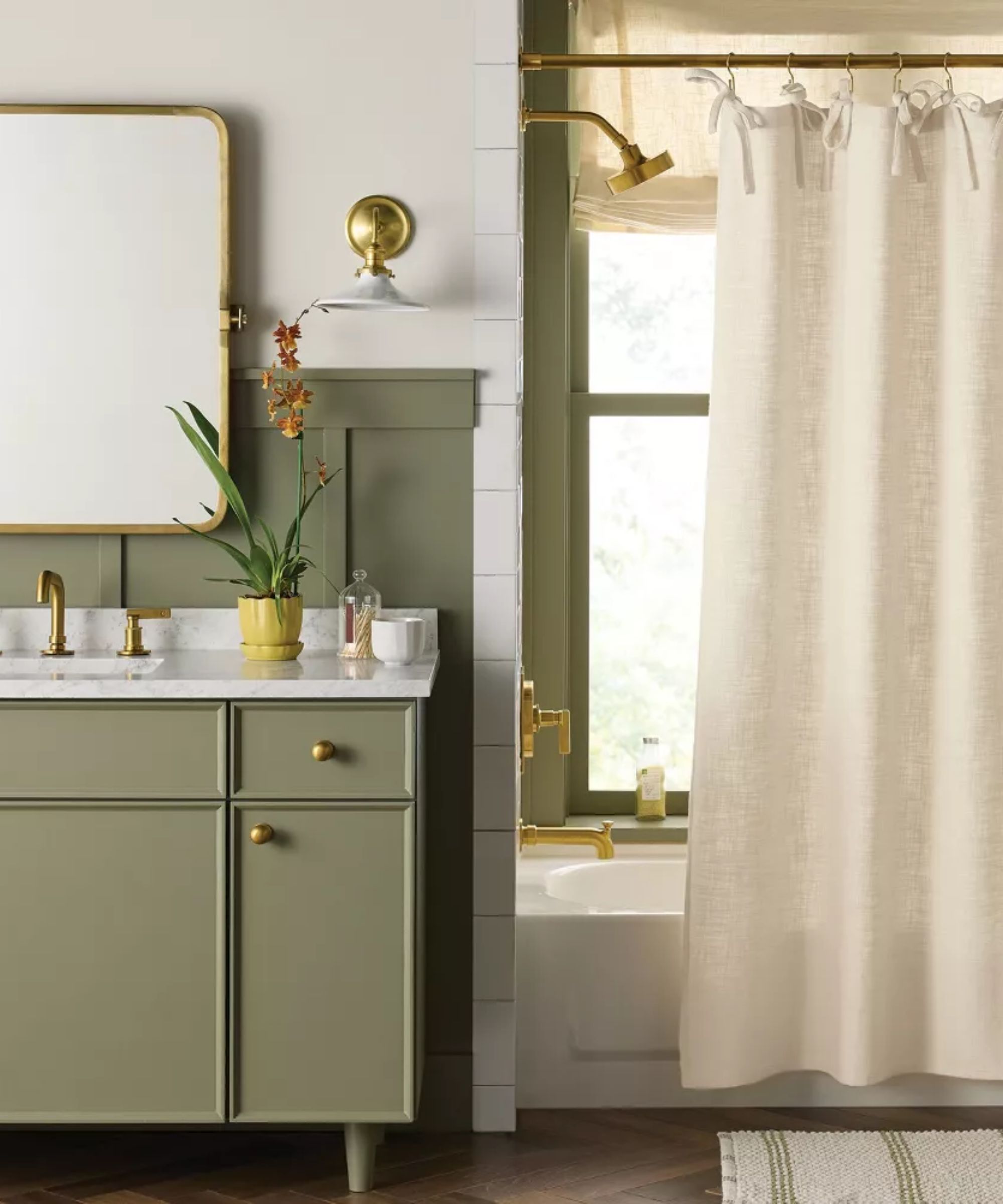 a bathroom with a soft green painted vanity with a marble counter surface, brass hardware, a plant on the counter, a mirror above, and a wall sconce to the right. Also to the right is a shower with a beige curtain with bows along the top