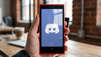 Hand holding a red Nokia Lumia smartphone displaying the Discord logo against a bright screen. Background shows a blurred room with large windows.