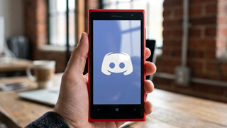 Hand holding a red Nokia Lumia smartphone displaying the Discord logo against a bright screen. Background shows a blurred room with large windows.