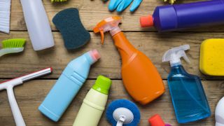 A range of cleaning products and tools on a wooden surface, including bleach, a squeegee, a sponge and spray bottles