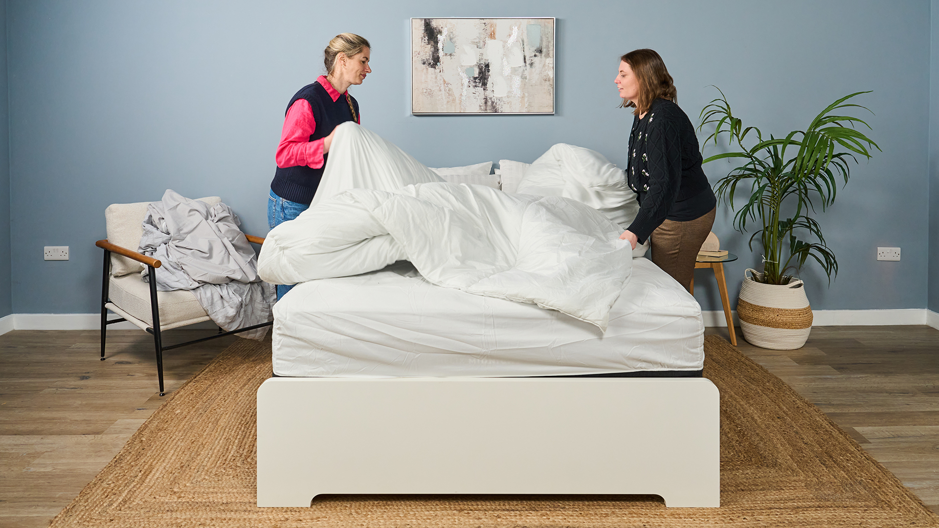 Two women stand either side of a mattress on a bed frame as they pull back the bedding