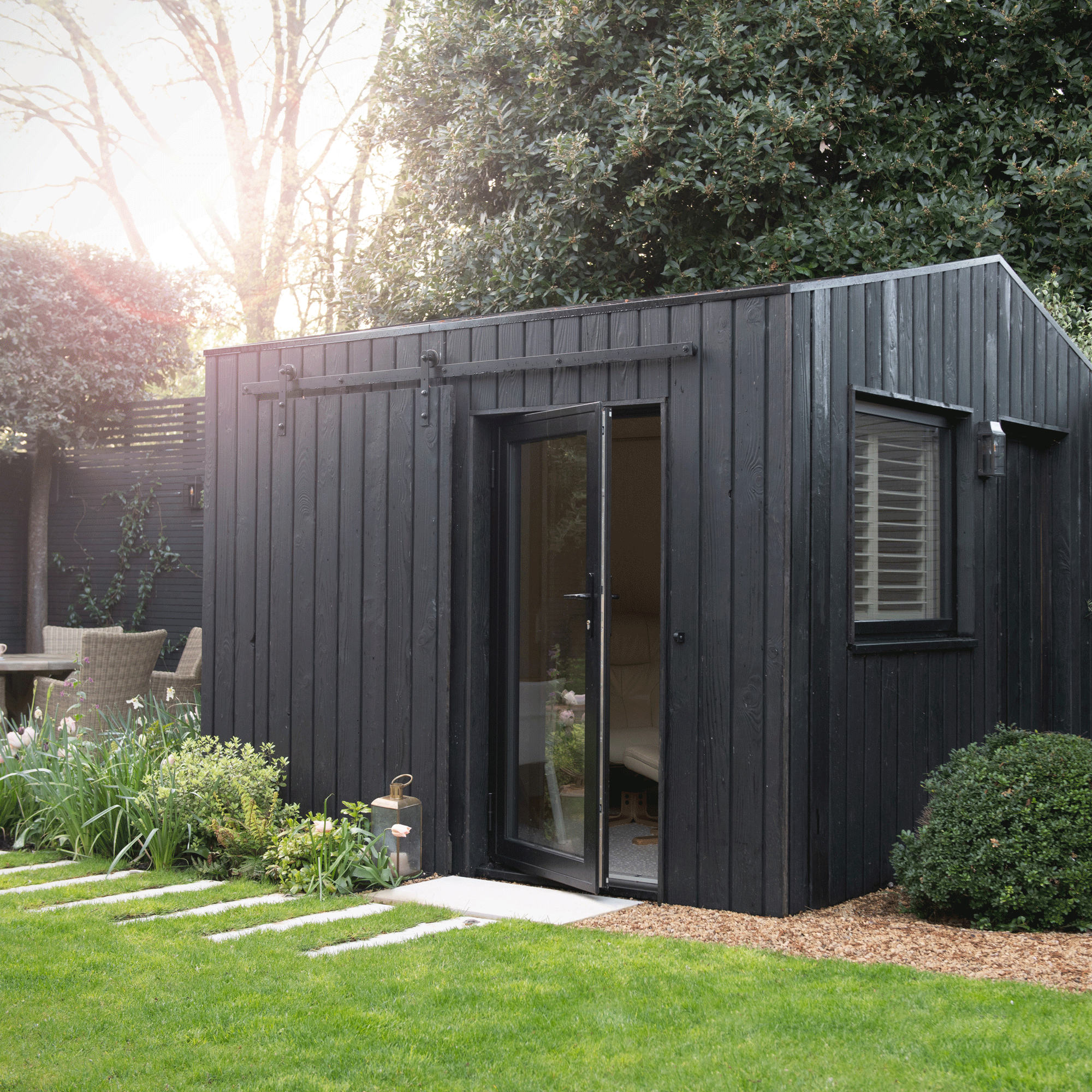 a garden building with black wood cladding beside a lawn and patio area with a dining set