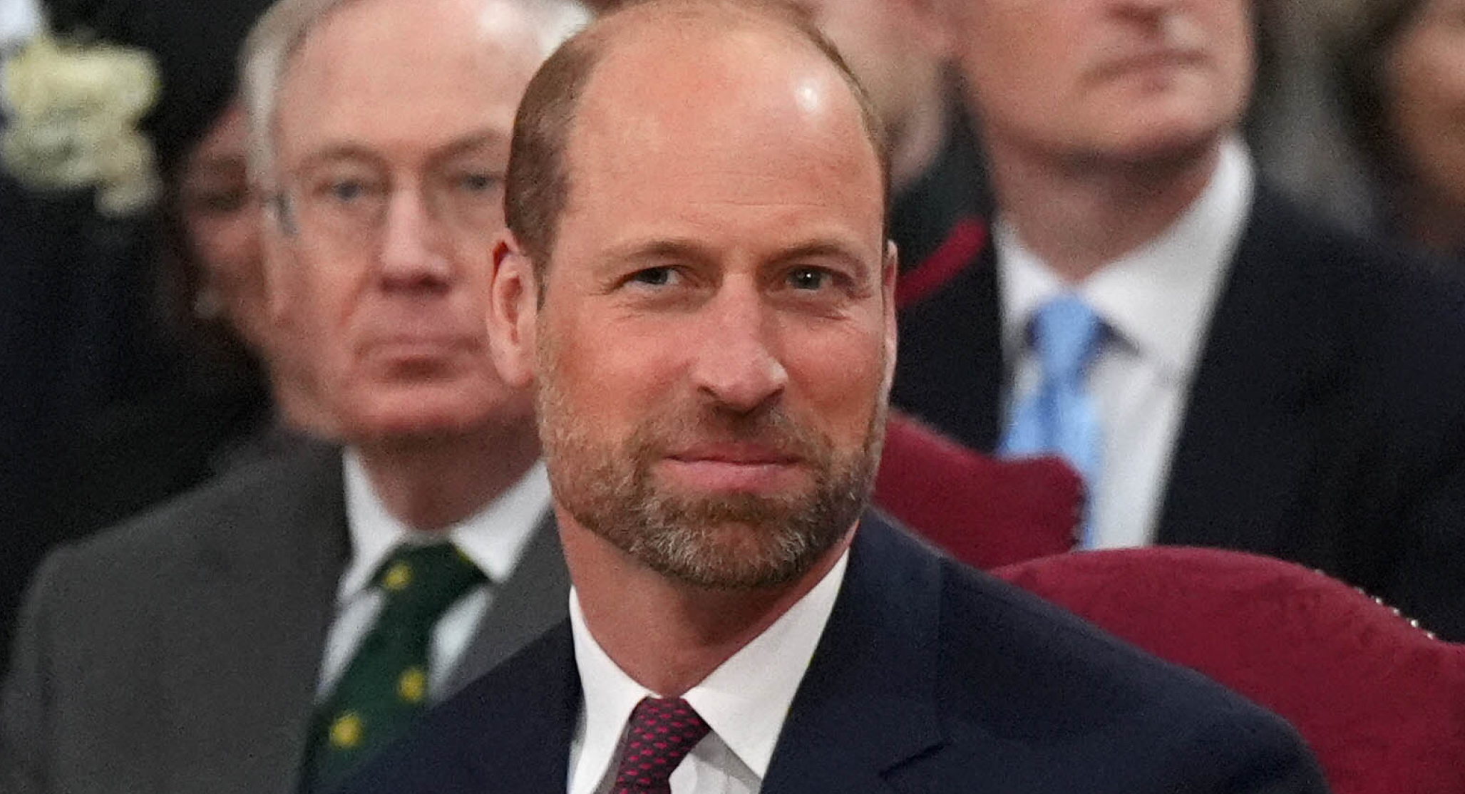 Prince William has a beard and a bald head and is smiling while wearing a navy suit and tie