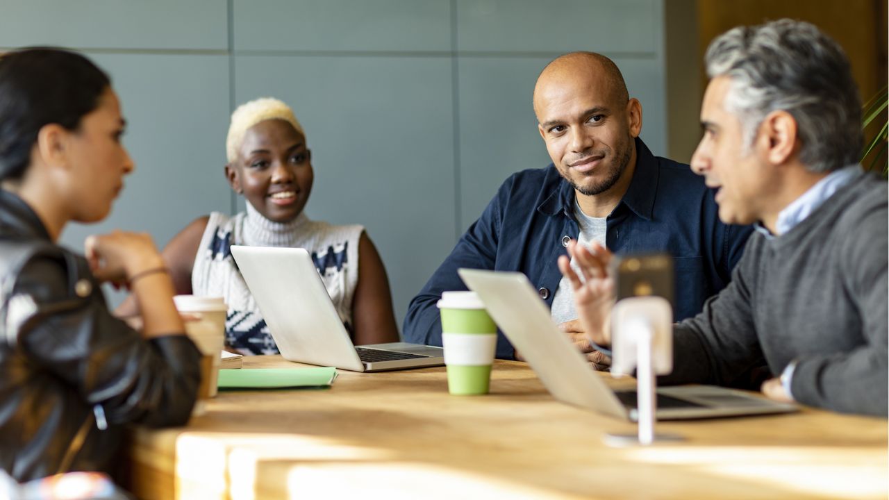 Four executives smile as they sit around a conference table with laptops in front of them.