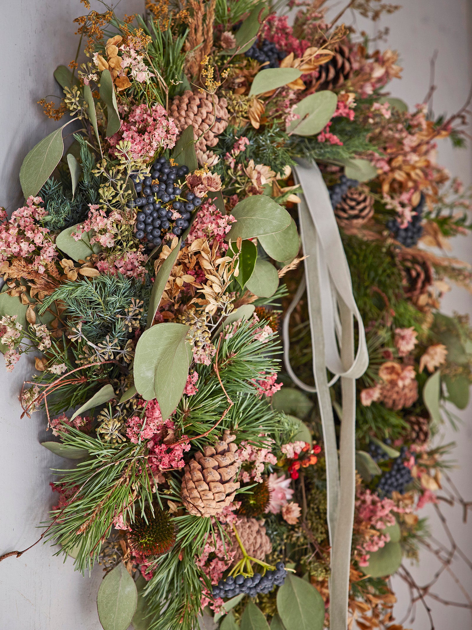 A Christmas wreath made of dried and fresh foliage, seed heads and pinecones, green leaves, black berries and silver ribbons.