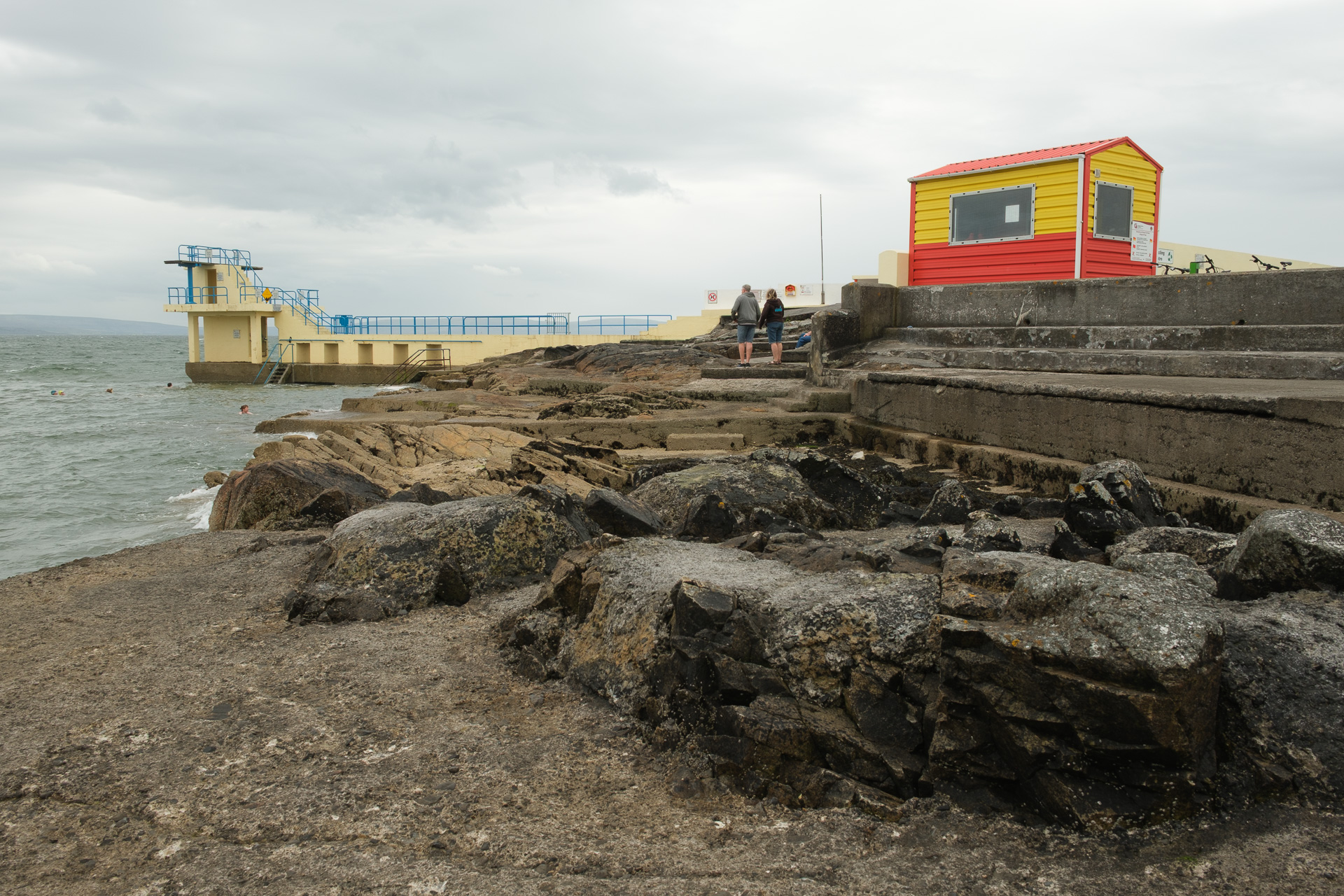 A rocky seafront with concrete steps, a red and yellow hut and a diving tower beneath cloudy skies.