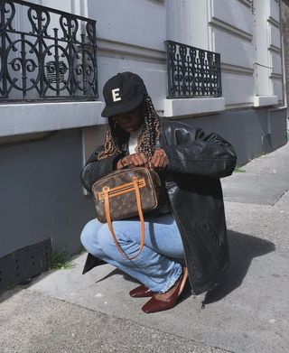 A influenciadora francesa Audie Julie em boné de beisebol, jeans, jaqueta de couro e bolsa com logotipo Louis Vuitton.