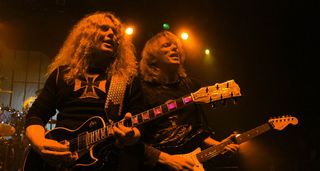 The late John Sykes plays his Les Paul Custom as Scott Gorham plays his Strat during a live Thin Lizzy performance in 2006. Both are bathed in red/orange stage lights.