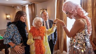 Fiona Dolman, in a floral dress and black feather boa as Sarah Barnaby, stands with Annette Badland, in a yellow top and orange boa as Fleur Perkins, as Felix Kai, in a pink wig and silver dress as Malik Payne, holds her hand and Neil Dudgeon, in a suit as DCI Barnaby, looks on in Midsomer Murders.