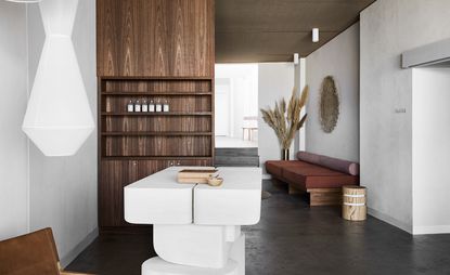 Interior view of the reception area at Studio Warrior One featuring white walls, grey floors, wooden shelving, a white sectional desk, seating with bolster cushions, dried plants and a geometric light fixture
