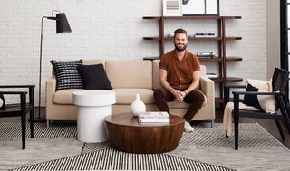 Bobby Berk sitting on a beige couch with his Karastan Rug, round timber coffee table and white brick wall