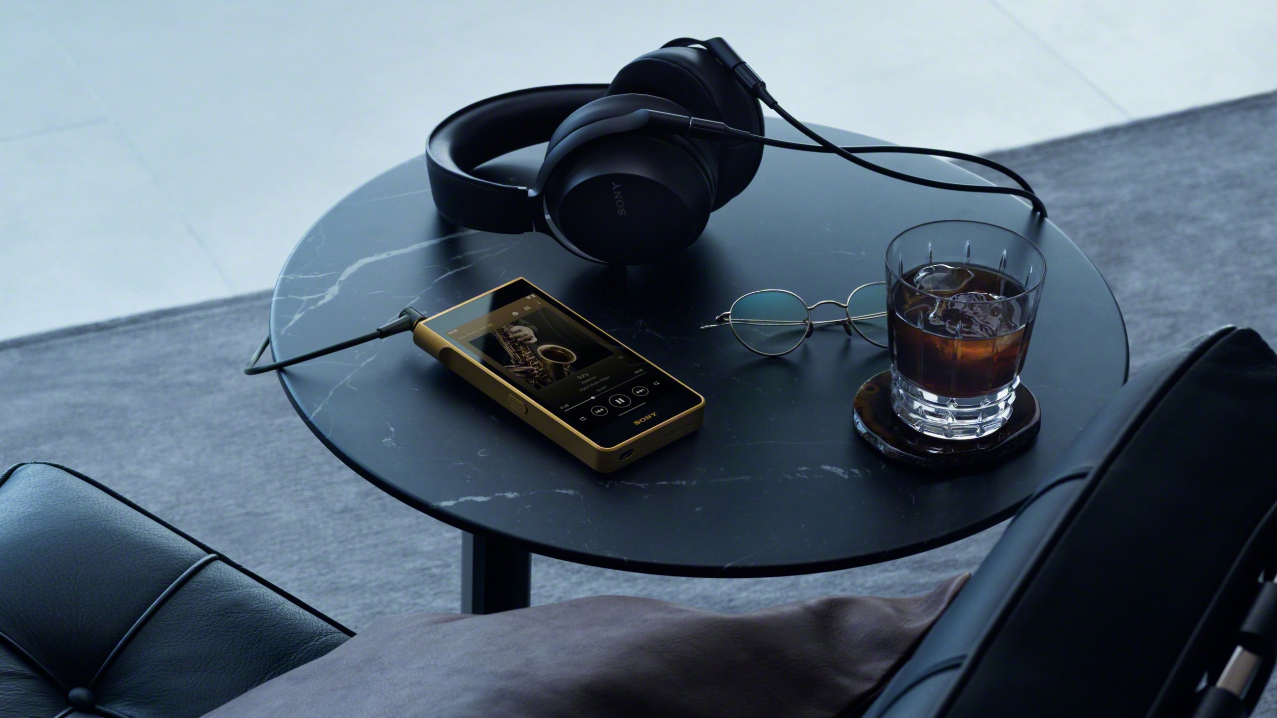 The Sony NW-WM1ZM3 Walkman on a table with a pair of over-ear headphones plugged into it. Also on the table are a dark drink with ice in a whisky tumbler and a pair of glasses.