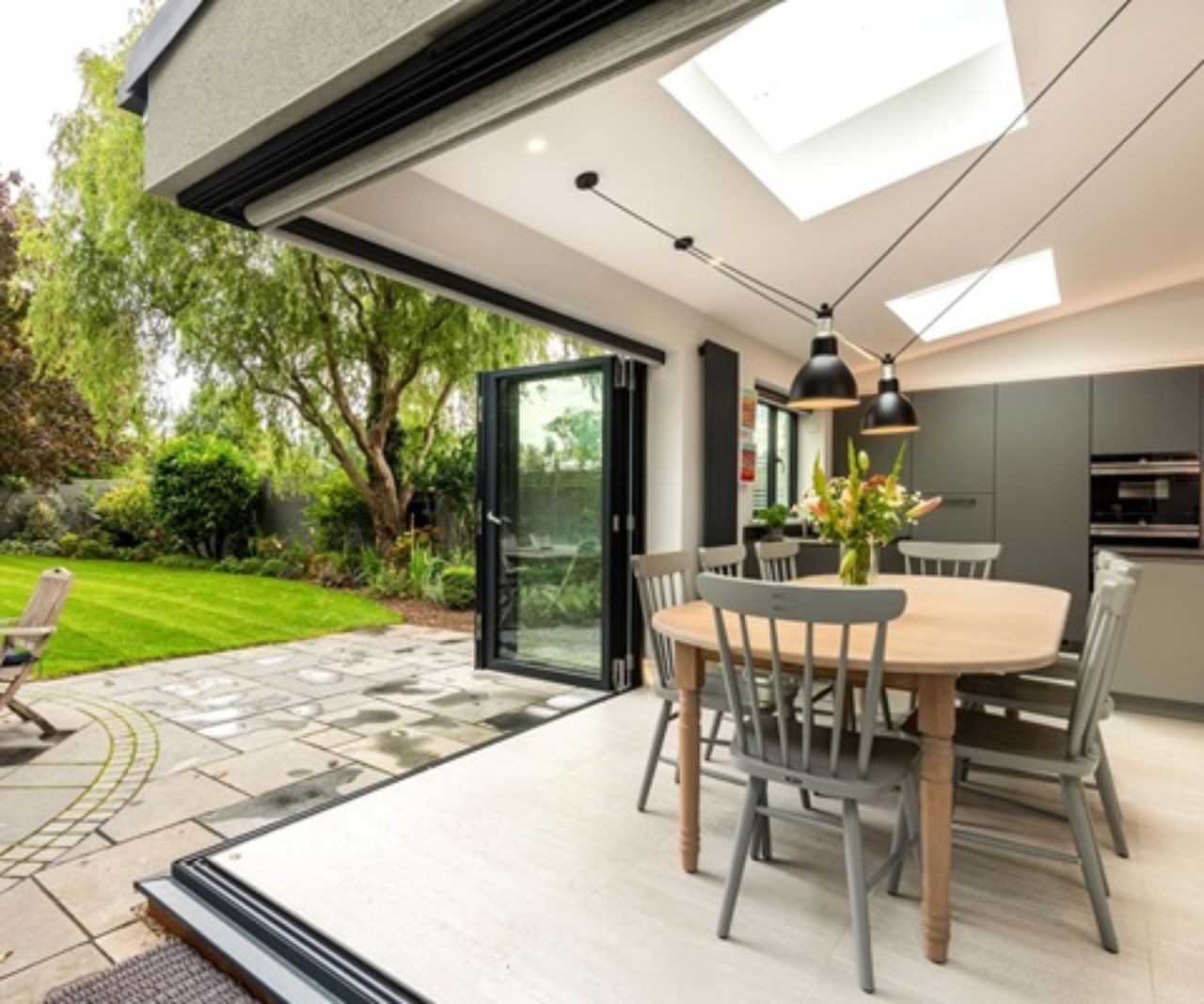 Corner of a kitchen diner extension with bifold doors from Kloeber peeled back to expose the interior to the garden. The kitchen features grey kitchen units; grey dining chairs have been paired to match the units.