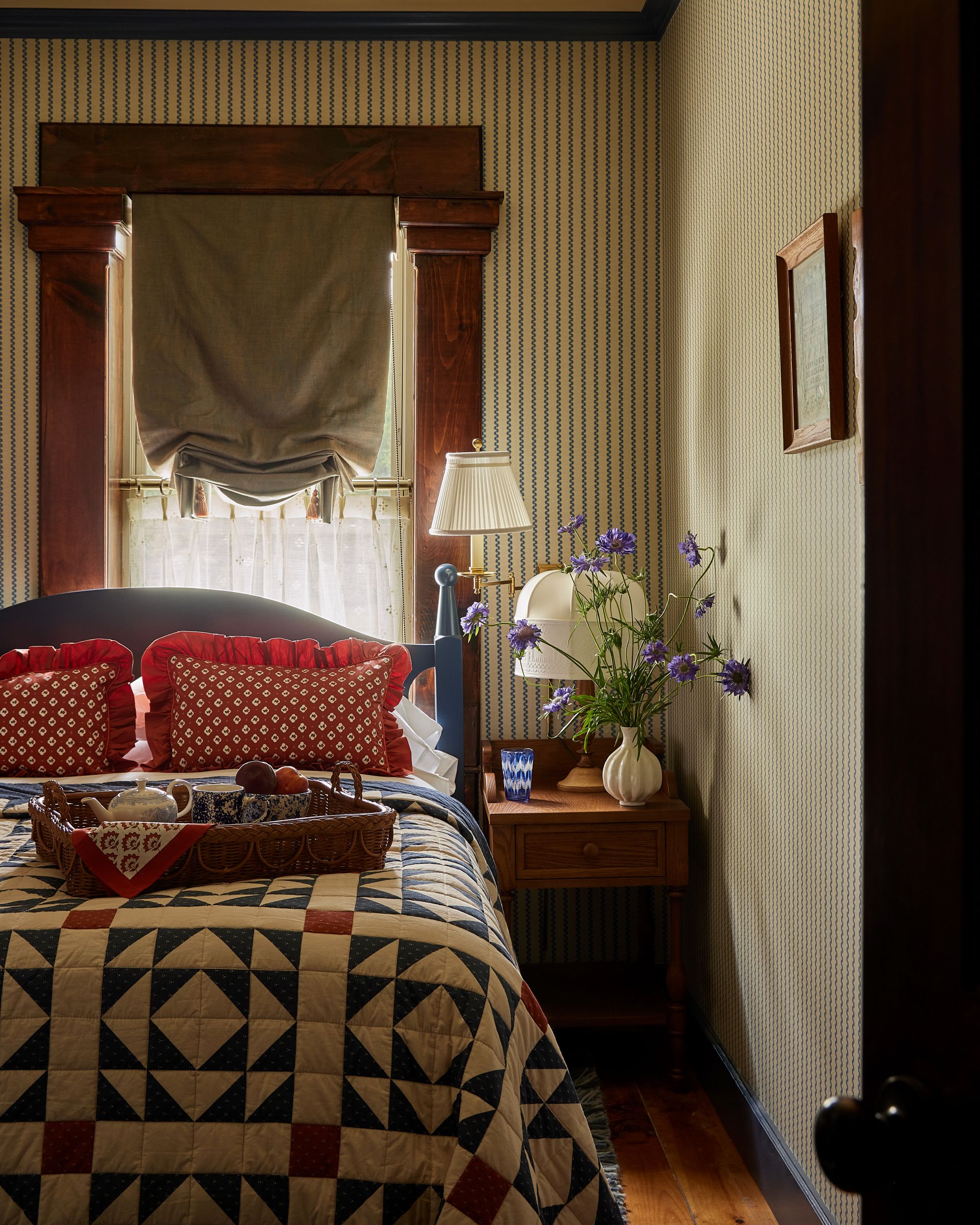 a bedroom with a patchwork bedspread, red pillows, striped wallpaper, and a floral arrangement on the side table