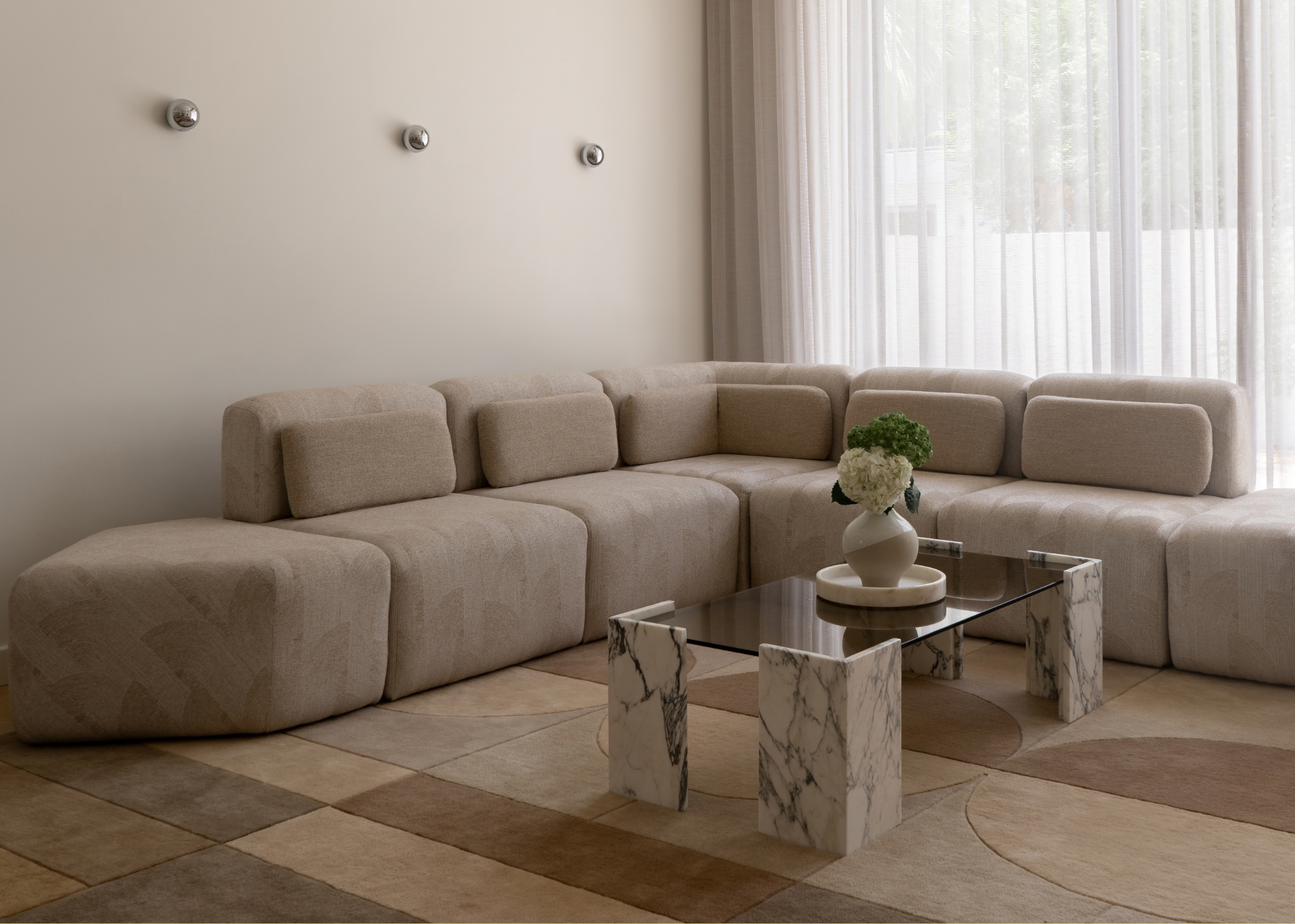 A minimalist suede sectional couch on a geometrically patterned neutral area rug with a marble and glass coffee table