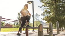 woman doing the step-up exercise onto bench in local park, wearing workout clothes and smiling in the sunshine under street light