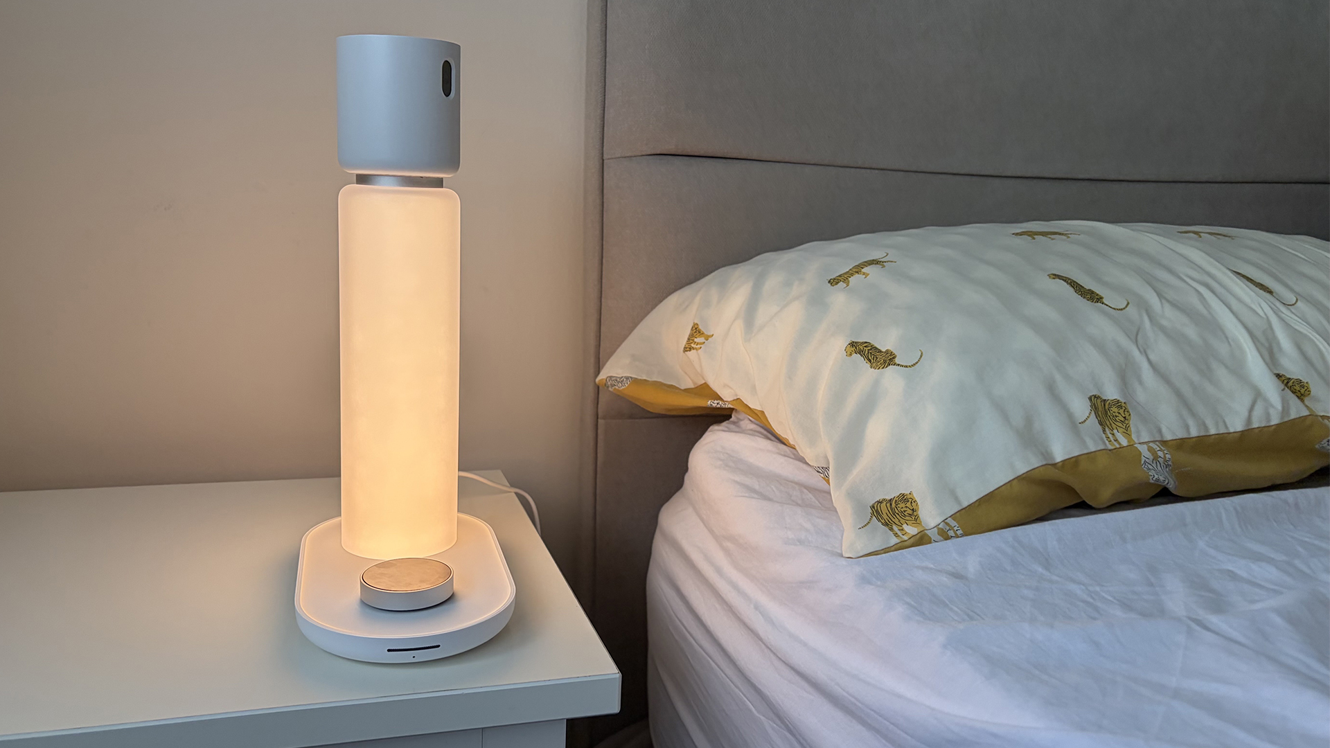 The Sleepal AI Lamp on the nightstand next to the bed in our reviewer's bedroom. The lamp is lit up with a warm, amber light