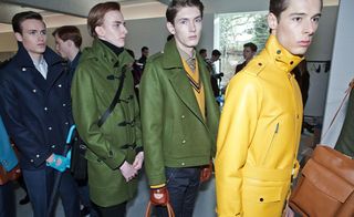 Four men in a line, part of the fashion show one with a yellow coat, one with a navy coat and two with green coats.