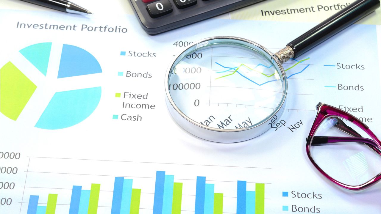 A magnifying glass, calculator and a pair of reading glasses sit on top of paperwork showing graphs about stocks and bonds.