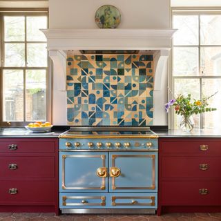 a pale blue range cooker with brass details beside dark red kitchen cabinetry and a splashback made from matching pale blue graphic tiles