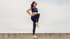 A woman stands with her hands on her hips and one leg raised to hip height. She wears a sporty crop top, leggings and sneakers.