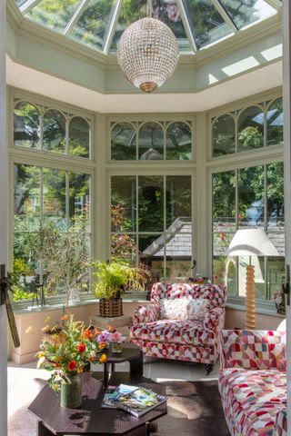 A charming conservatory with sage green framing, a glass roof, and arched windows. Patterned armchairs, lush greenery, and a sparkling chandelier create a cosy yet elegant retreat.