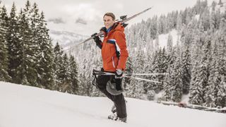 A skier in a red jacket carries his ski gear uphill