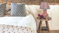 A white and wood bedroom with double bed with patterned quilt, cushions, and lamp on a circular wooden nighstand.