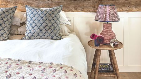 A white and wood bedroom with double bed with patterned quilt, cushions, and lamp on a circular wooden nighstand.