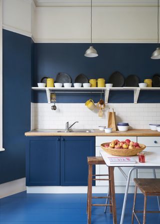 inky blue kitchen with walls and cabinetry in navy, white cabinets and table, white tiled backsplash, open shelving, bright blue painted floor, white skirting