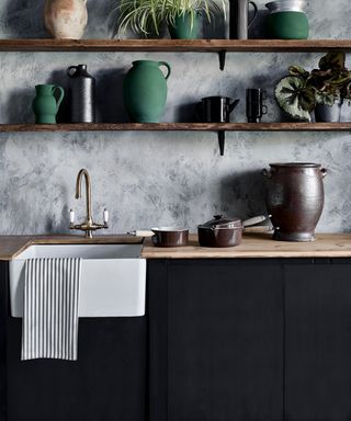 black kitchen cabinets with open shelving, wooden worktop, green vases, plants, brass faucet, white butler sink, grey painted wall,