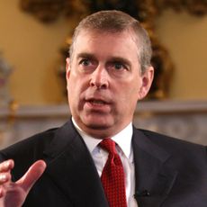 Ex-Prince Andrew wears a suit with a white shirt and a red tie and holds his hands in the air
