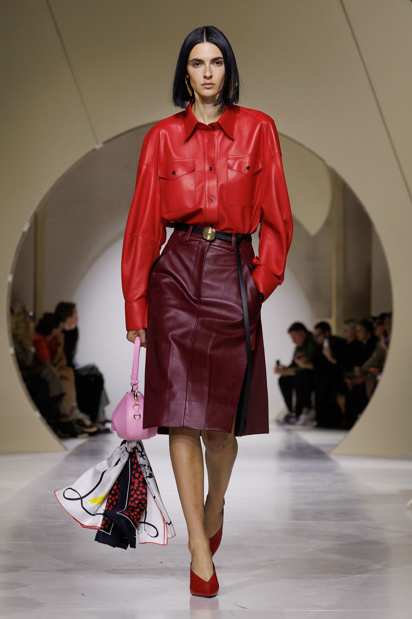 A Givenchy spring/summer 2027 runway model wearing a tomato red leather shirt tucked into a burgundy red leather skirt worn with red pumps.