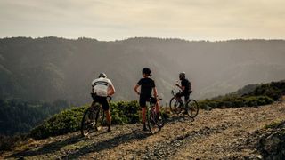 Three gravel cyclists - two male, one female - resting at the top of a gravel climb and looking into the distance