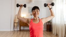 woman facing the camera smiling holding a black dumbbell in each hand to the sides of her head. she's wearing a pink vest and standing in a living room setting. 