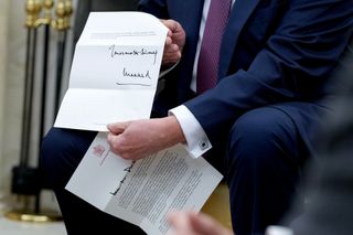 Donald Trump sitting in a chair wearing a blue suit and holding a letter up to the camera from King Charles