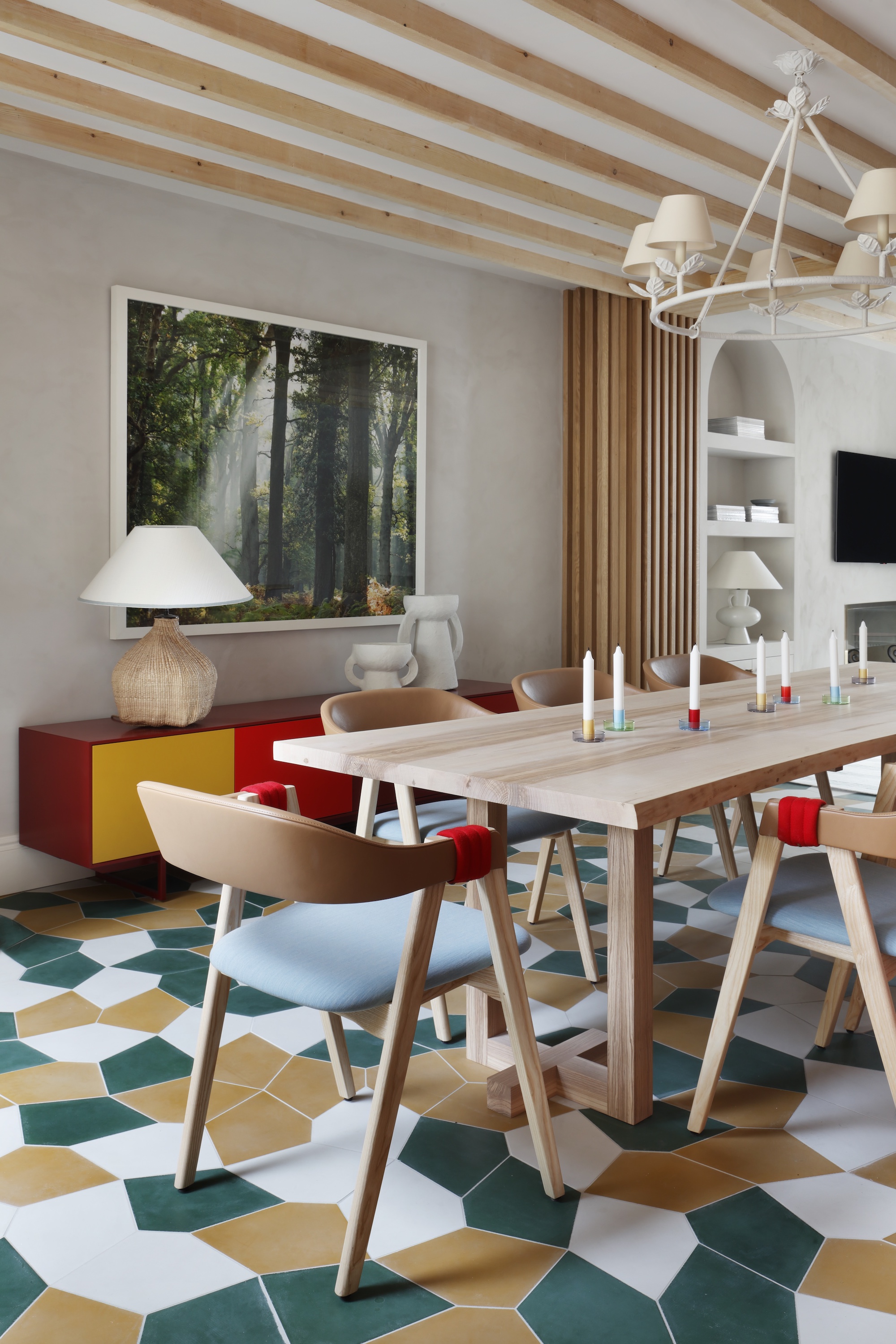 Dining room with a pentagon tiled floor in white, green and yellow. The dining table sits centrally with a cream chandelier above and a multicoloured credenza behind.