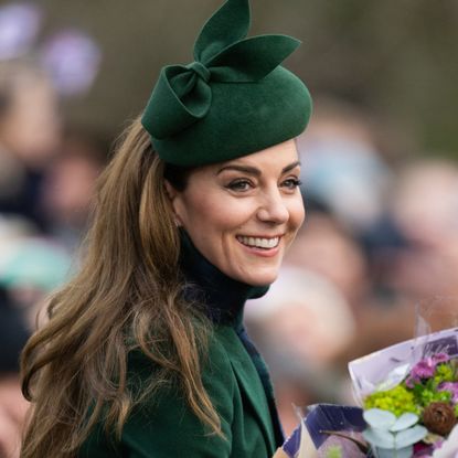 Kate Middleton wearing a green hat and coat smiling and holding flowers with a crowd of people blurred behind her