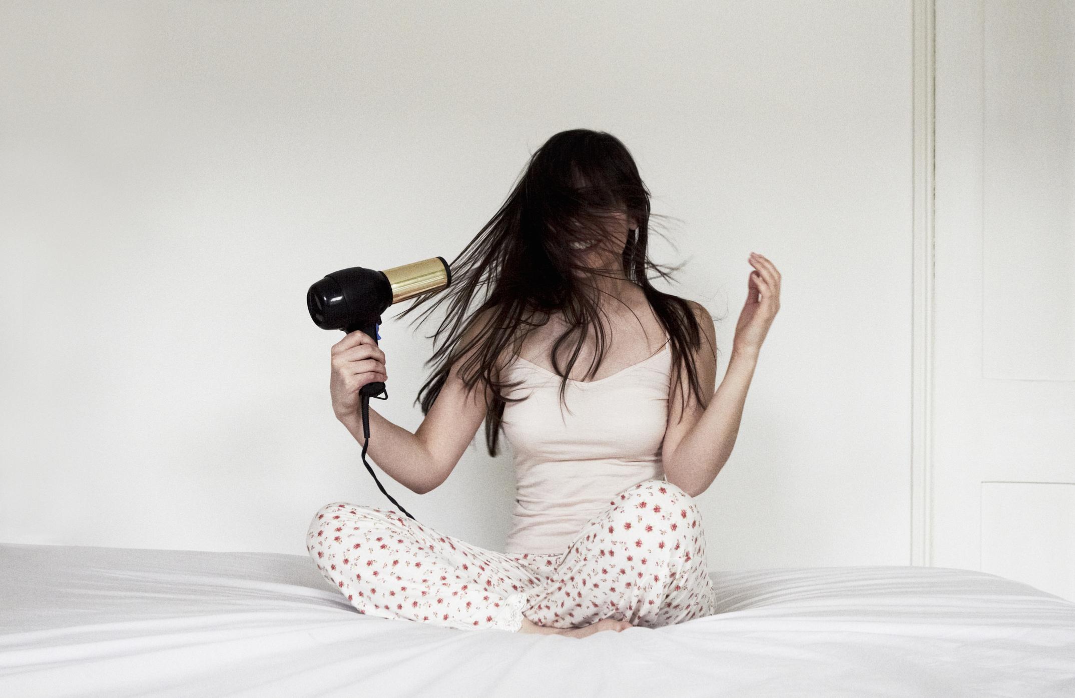  A woman sitting on a bed blow drying her long hair. 