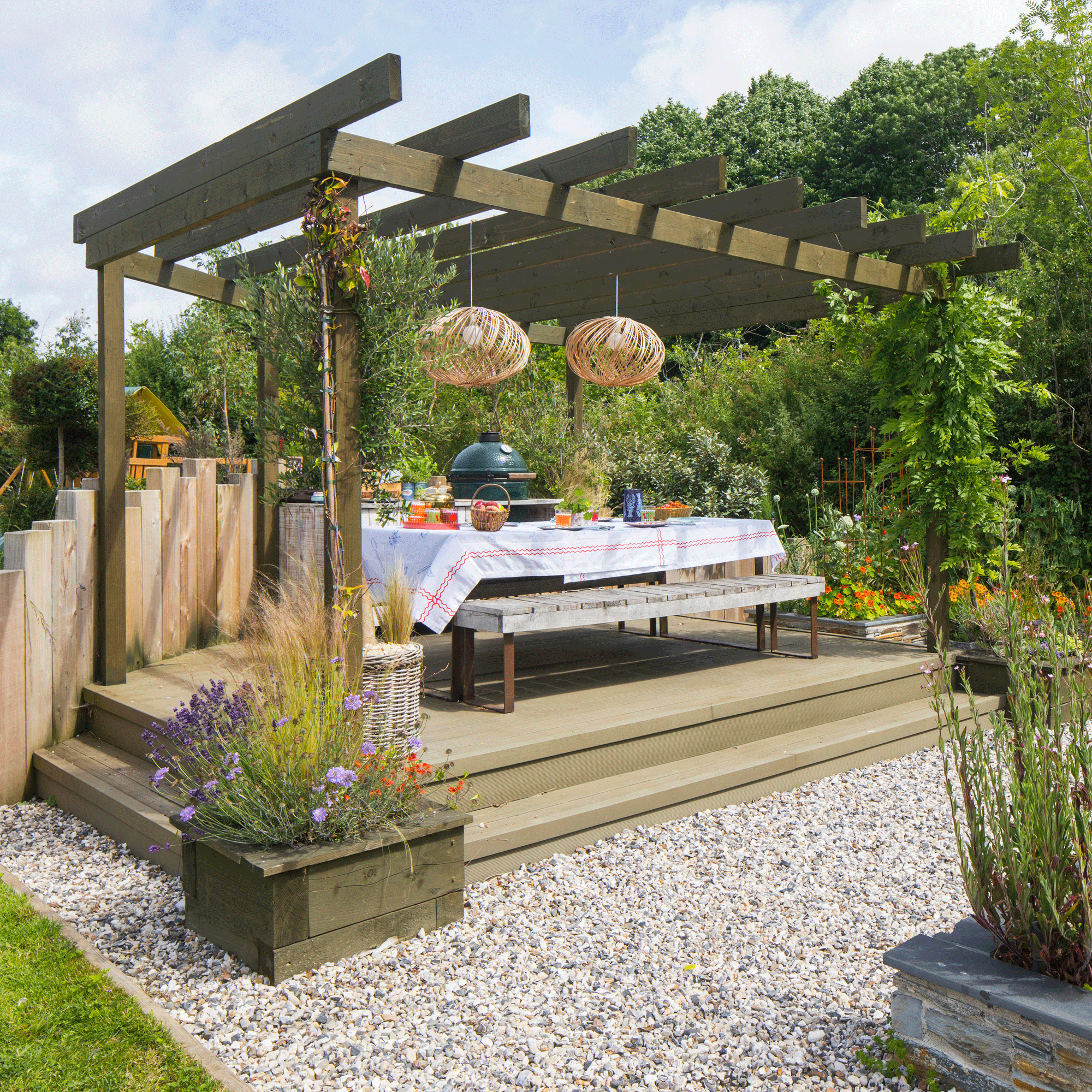 a garden with a raised decked area with a pergola above and a Big Green Egg BBQ and dining table and bench all set for a meal